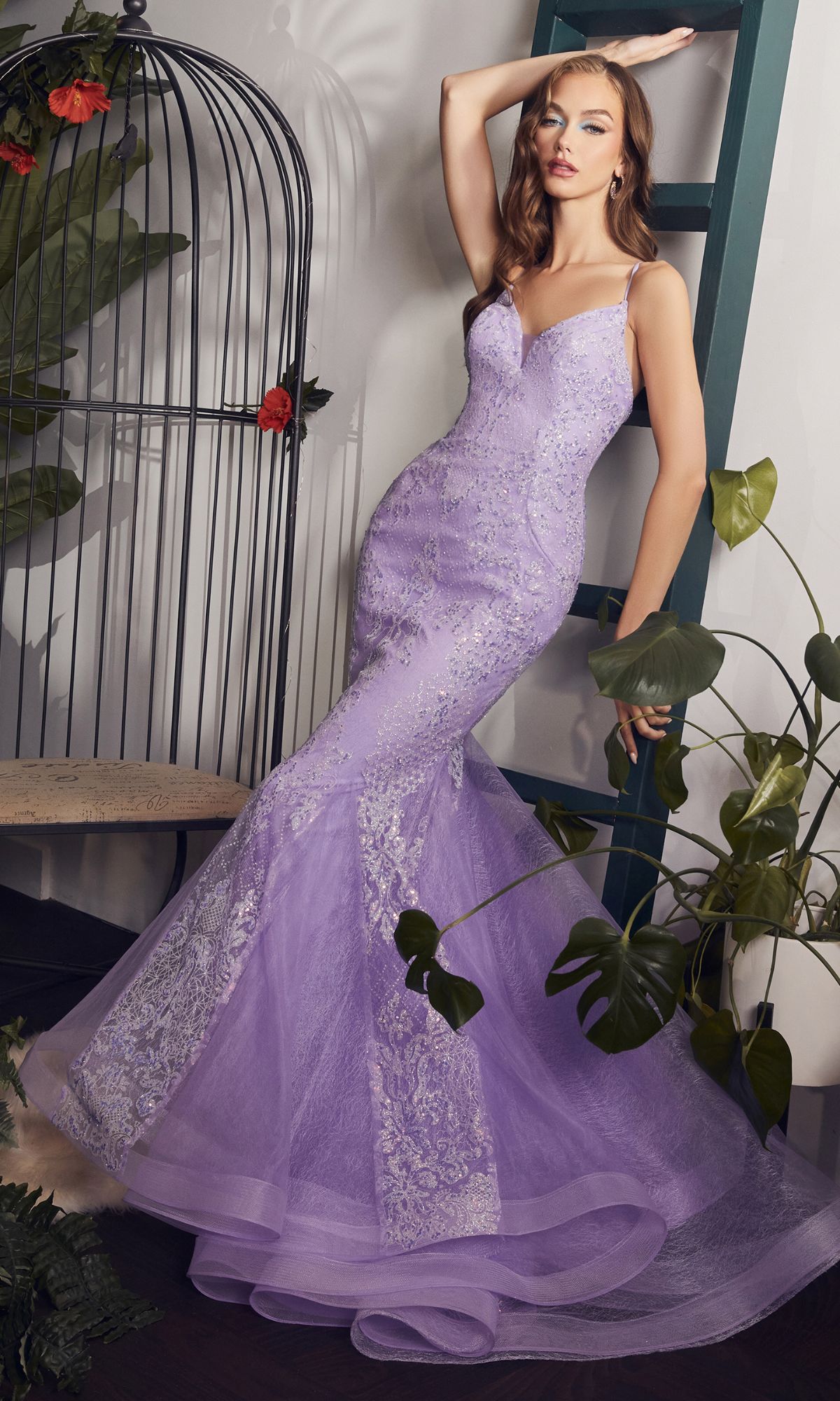 A woman poses indoors in the Strappy-Back Long Beaded Mermaid Prom Dress CC2279 near a green ladder and birdcage, surrounded by lush plants.